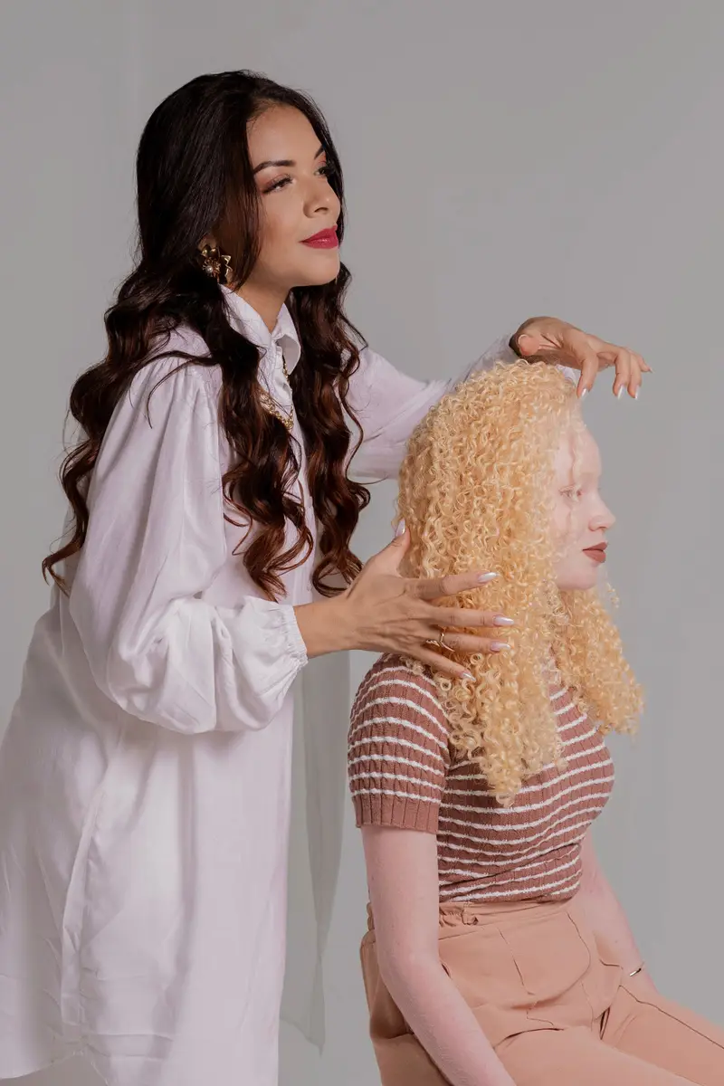 A hairdresser in a white shirt arranges the long blonde curls of a seated woman against a pale grey backdrop.